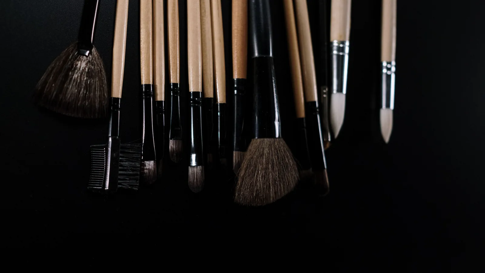 A selection of make-up brushes with wooden and black handles against a dark background.
