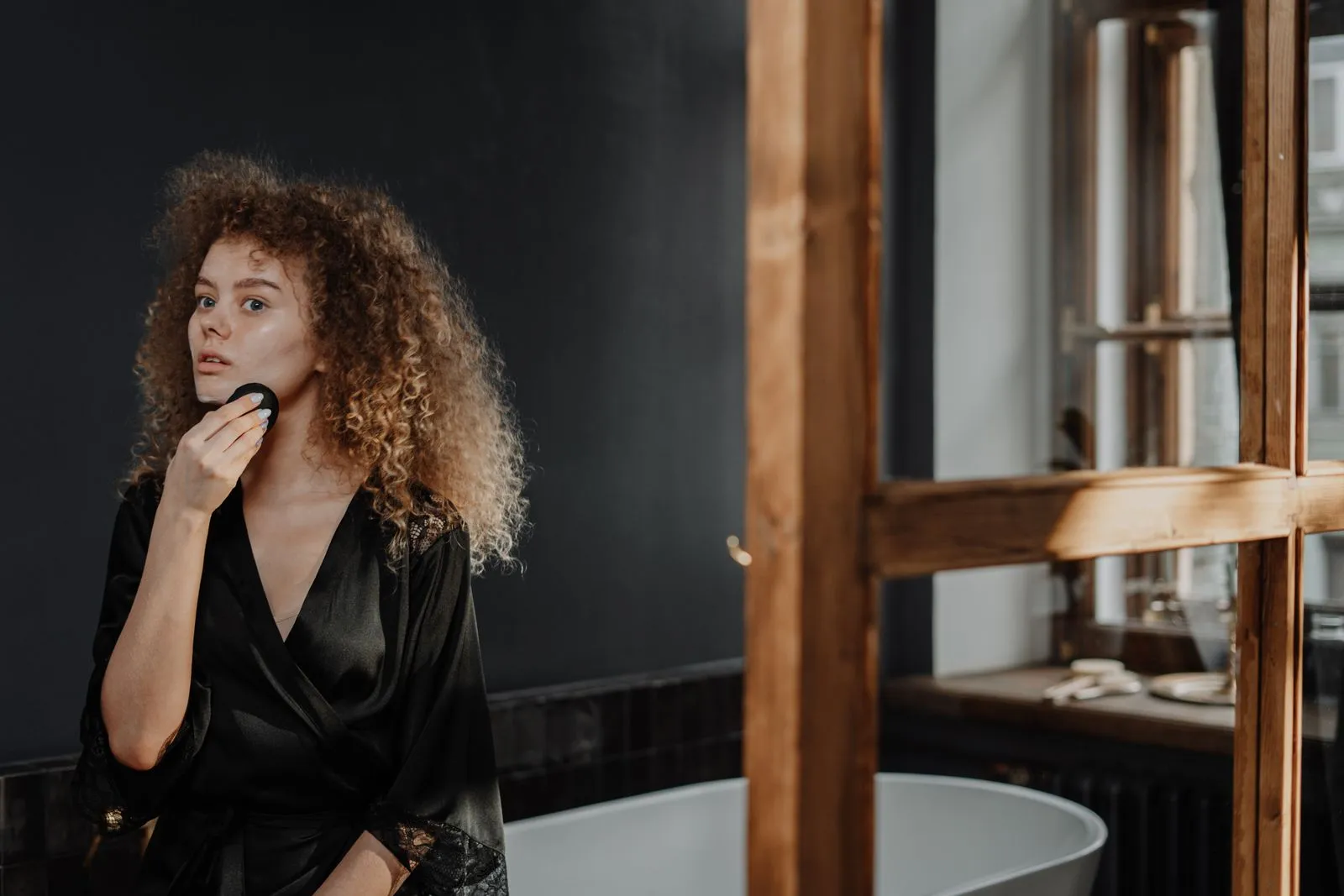 A young woman with curly hair does her beauty routine in a stylish bathroom, applying makeup.
