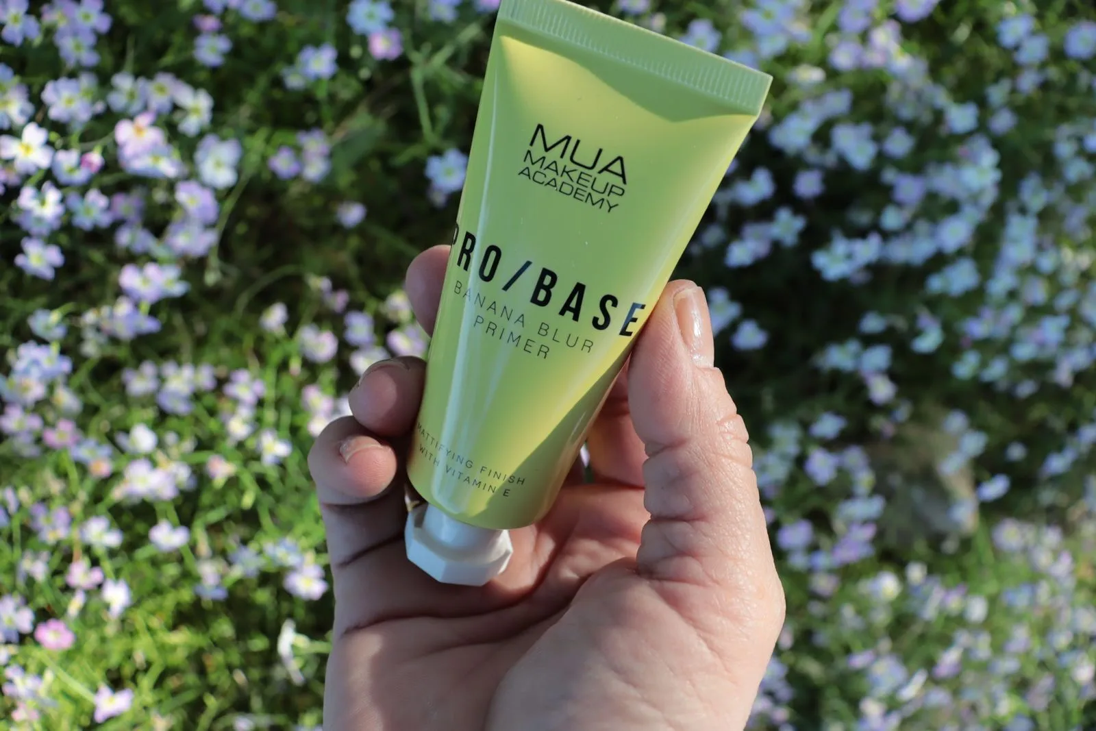 Close-up of a hand holding a makeup primer tube with flowers in the background