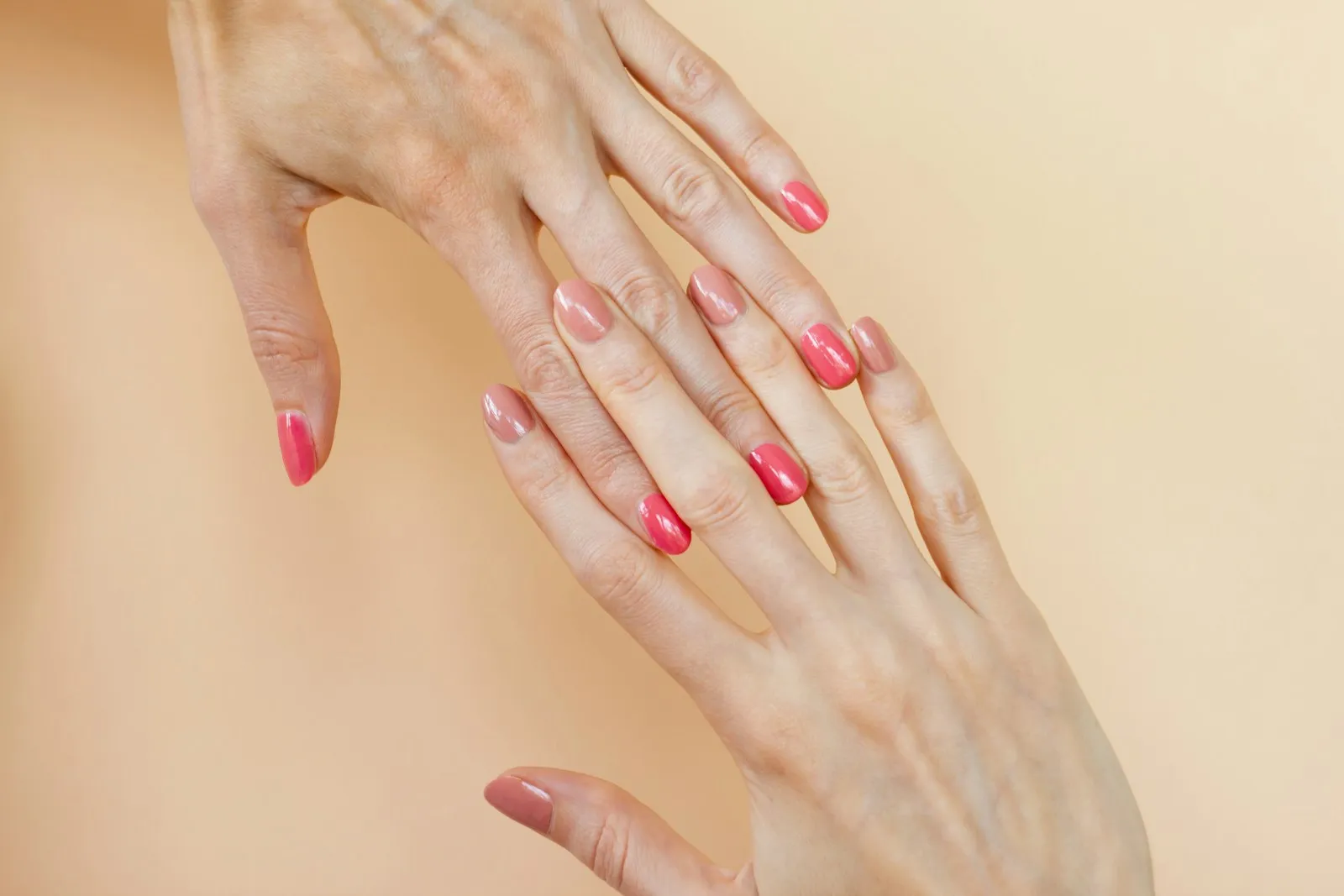 Against a warm sand-colored background are two hands with long fingers, the nails of which are painted in two different