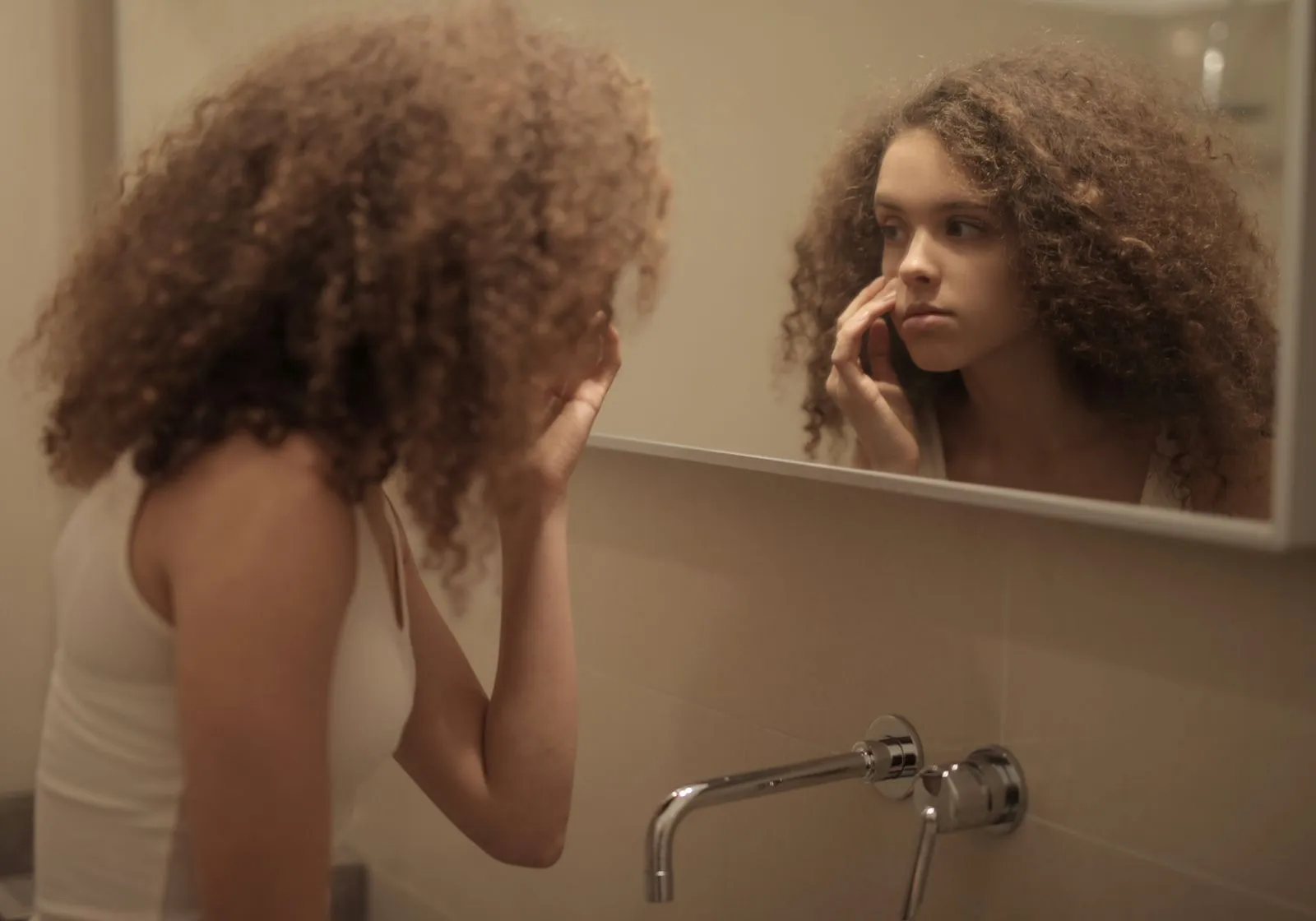 Teenage girl with curly hair examines her face in the bathroom mirror, reflecting contemplation.