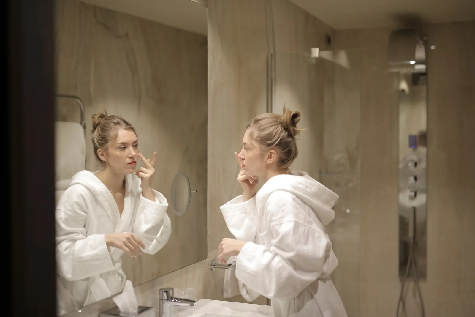 A woman in a white bathrobe applies moisturizer while looking at her reflection in the bathroom mirror.
