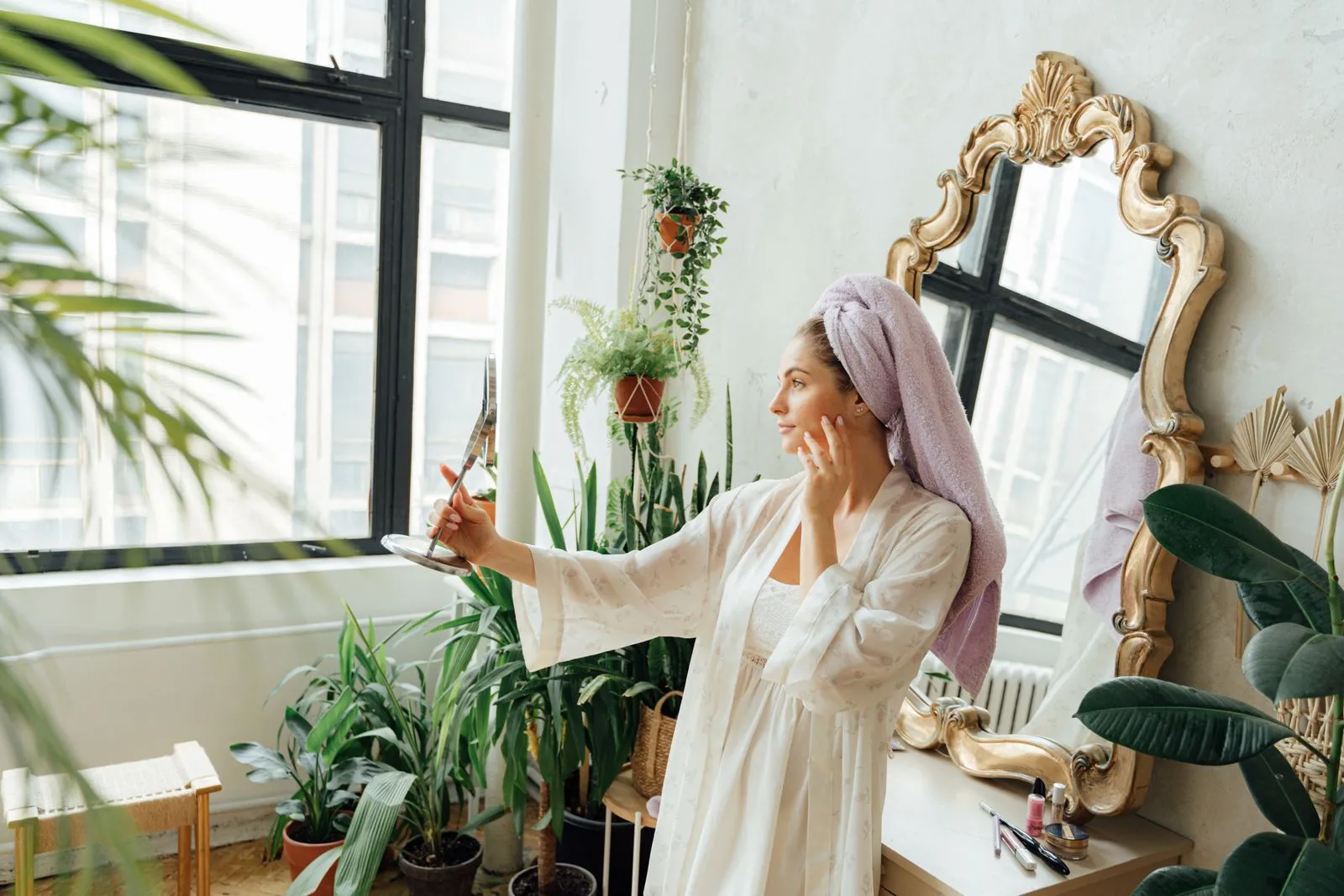 Adult woman in bathrobe and head towel applying skincare indoors, surrounded by plants.