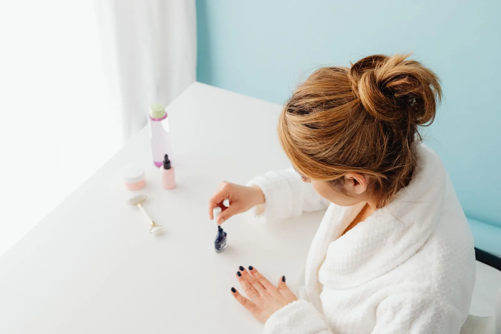 Woman in white bathrobe carefully painting her nails indoors, emphasizing self-care.