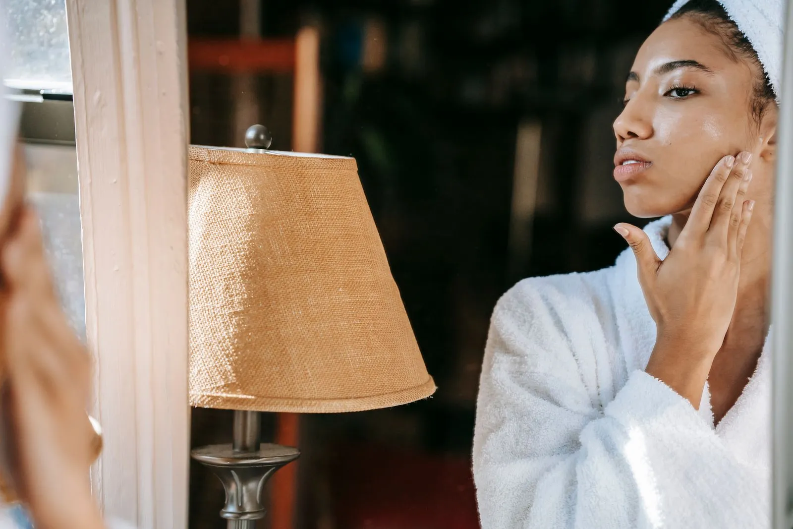 Crop young feminine ethnic lady in robe touching face while looking in mirror in house