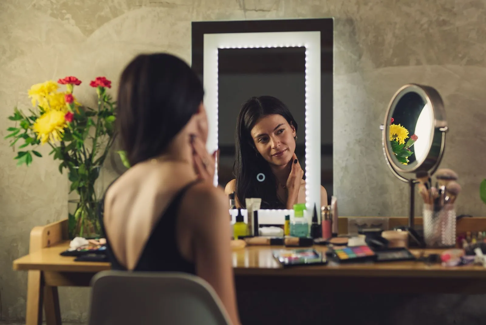 A woman sitting at a vanity, applying makeup before a mirror with reflection.