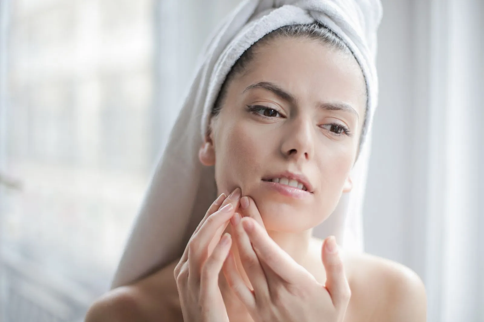 Young woman with towel examining her face during skincare routine in a bathroom setting.