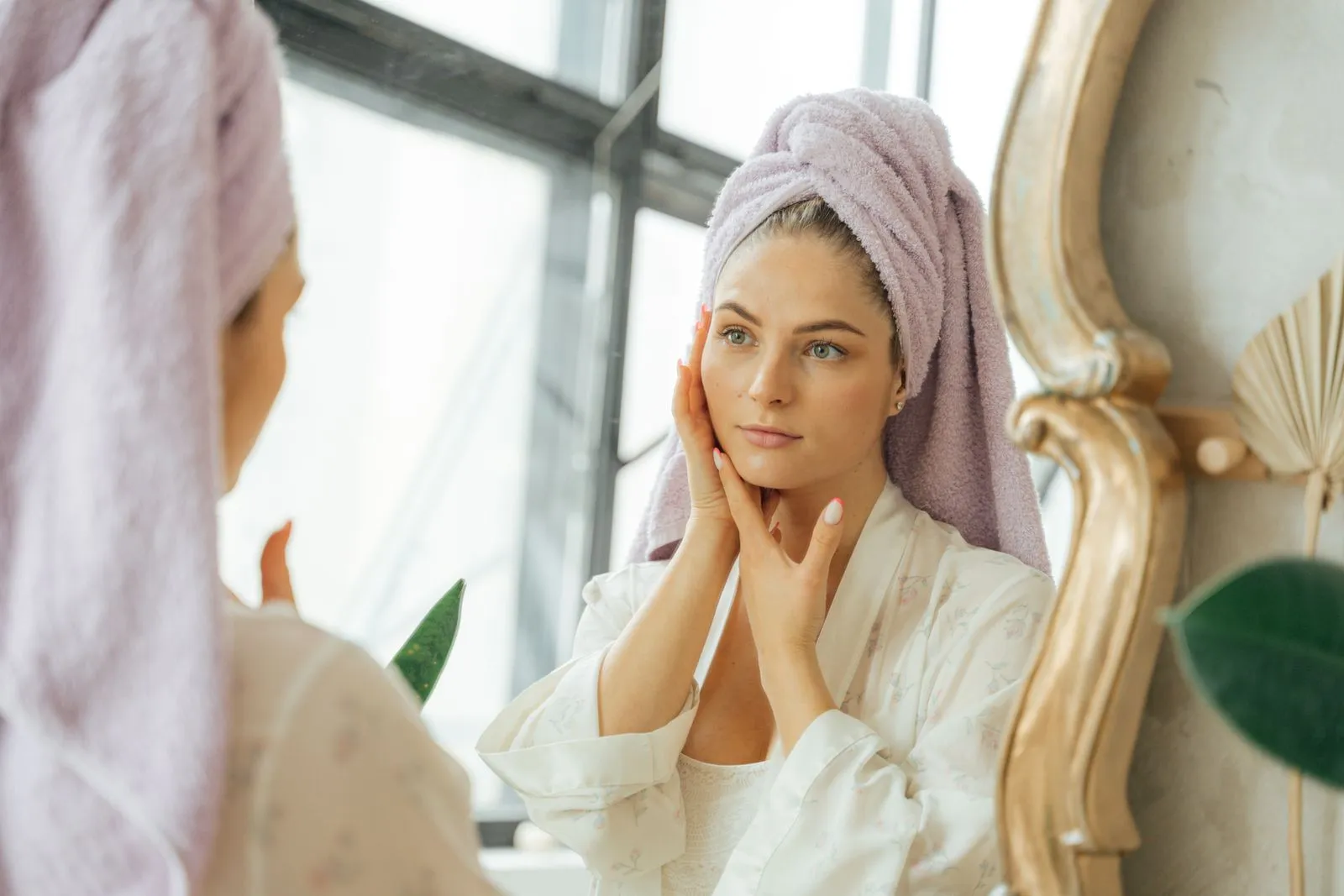 Woman in bathrobe with head towel touching face, enjoying skincare routine in front of mirror.