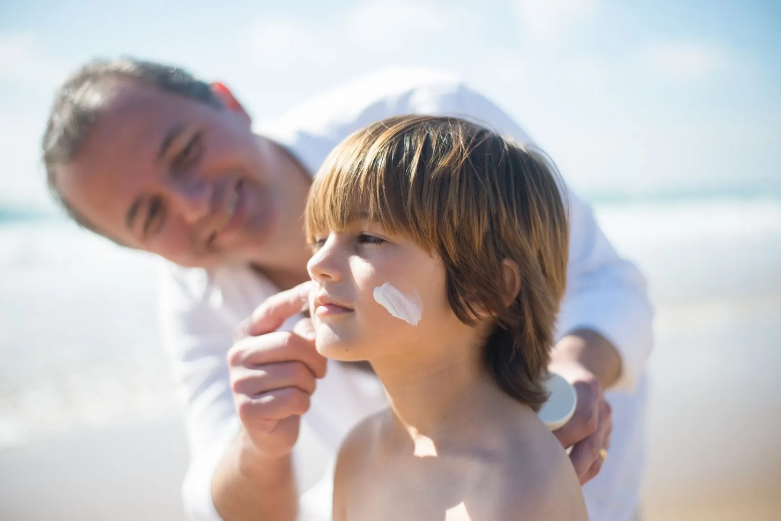 A father gently applies sunscreen to his son's face on a sunny day at the beach, ensuring skin protection.