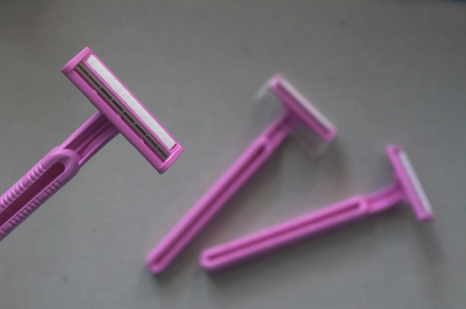 Three pink single-use double blade women's disposable razors with moisture strips for sensitive skin on grey background.