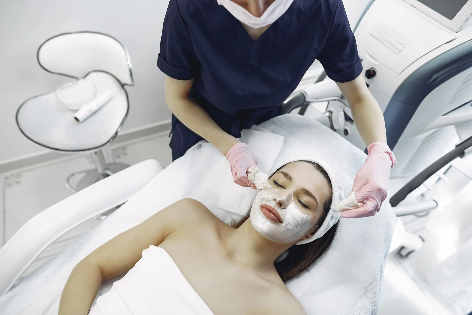 From above of dermatologist in medical coat and latex gloves doing face cleansing and wiping mask from face of female ly