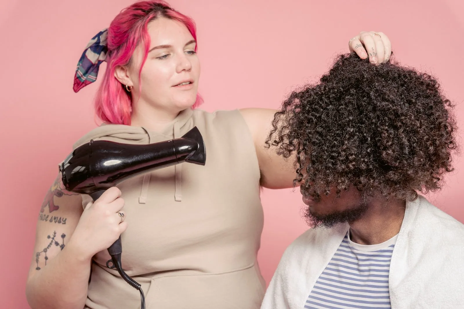 Female hairdresser with dyed hair drying hair of African American male model with Afro hairstyle against pink background