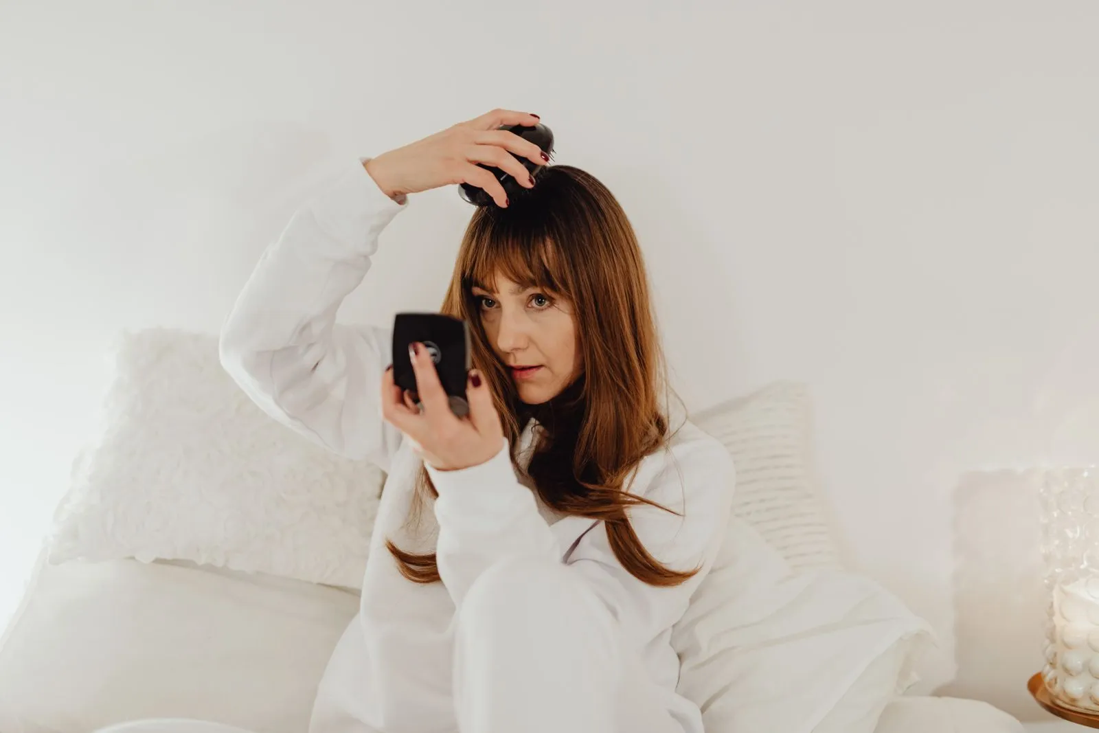 Woman in white attire combs her hair while seated on a bed with soft lighting.
