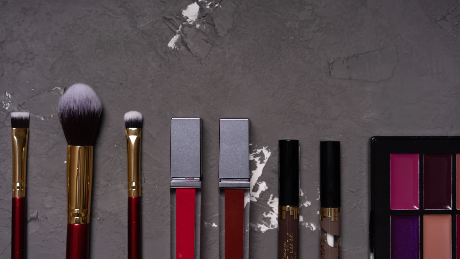 Make-up brushes, lip colours and a palette arranged on a grey surface.