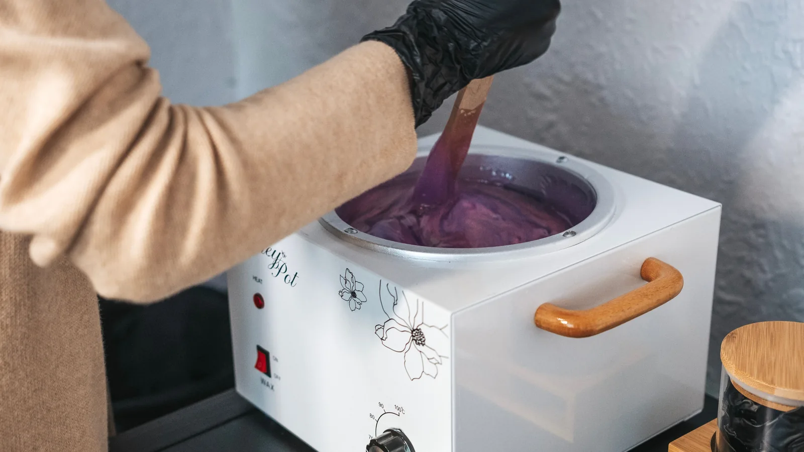 A black-gloved hand stirring purple wax in a white wax heater.