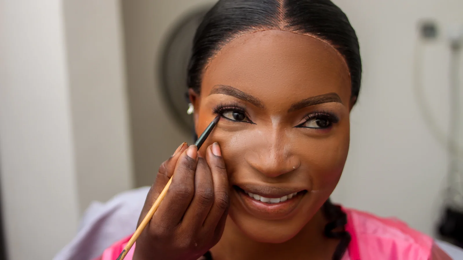 A person in a pink robe having eye make-up applied with a small brush.