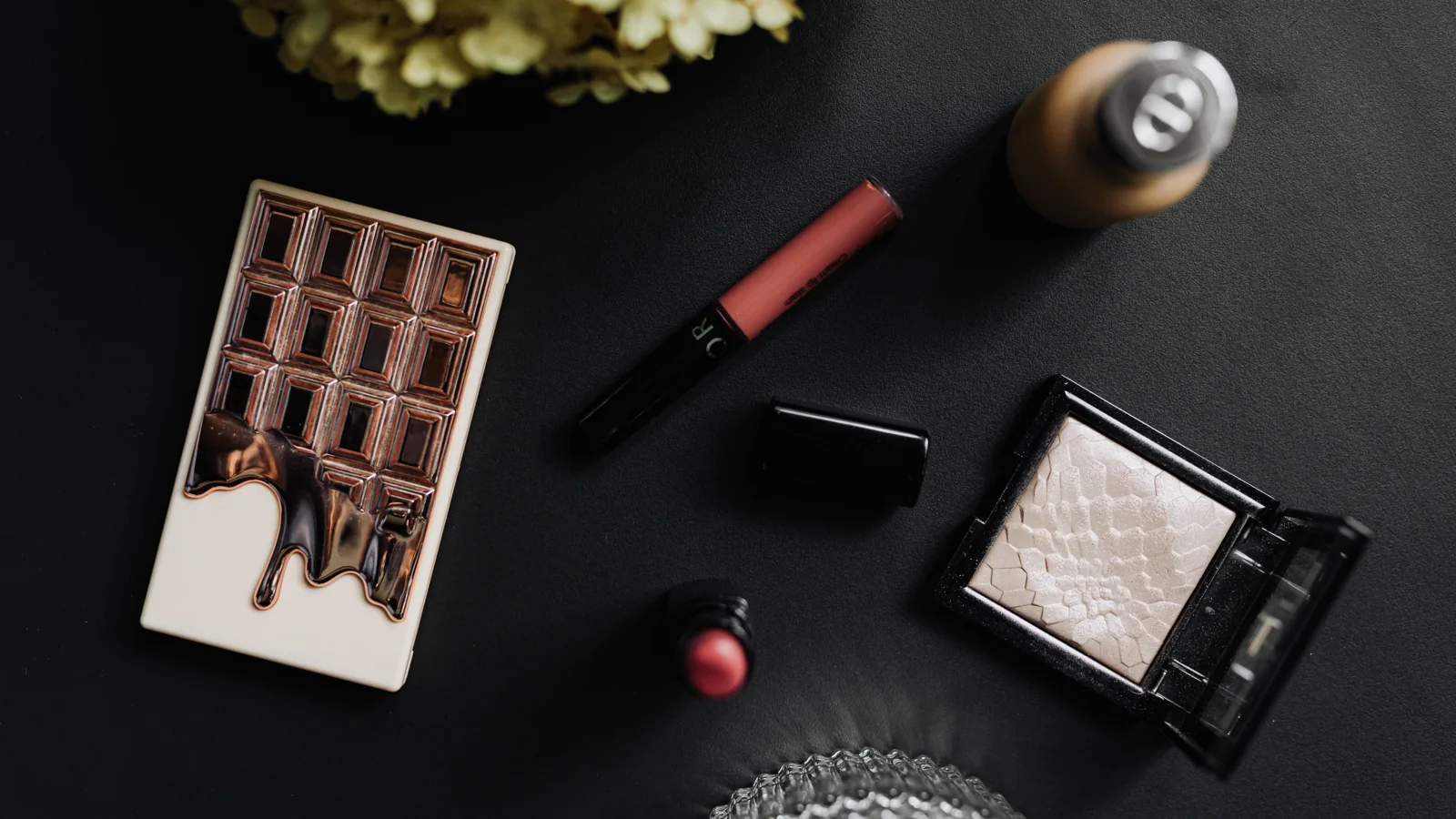 An eyeshadow palette, lip colour and other cosmetic items arranged on a dark table.