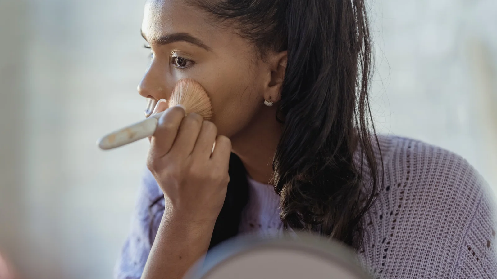 A person applying make-up to their cheek with a brush beside a mirror.