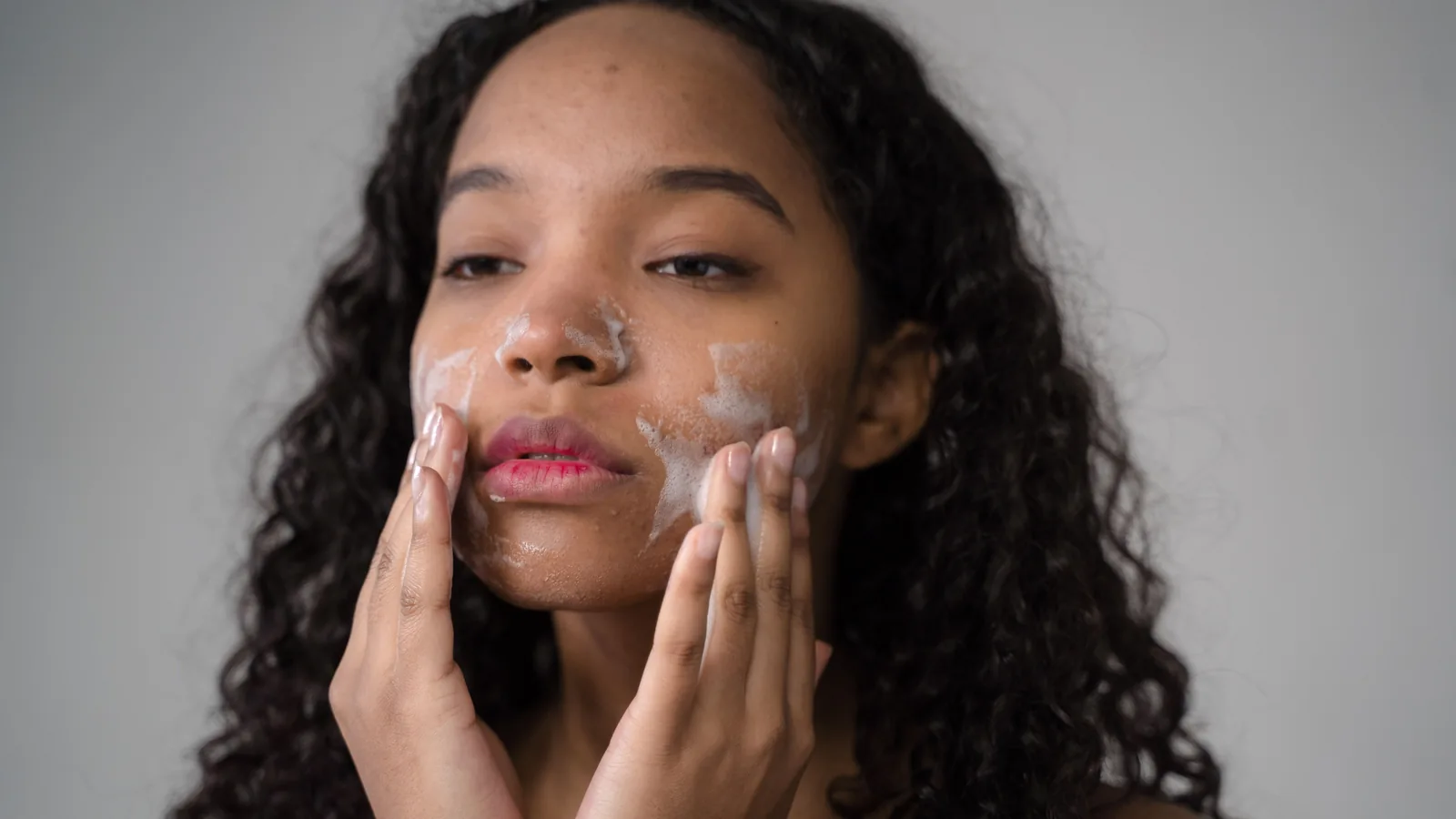 A person with curly dark hair washing their face with foam on their cheeks.
