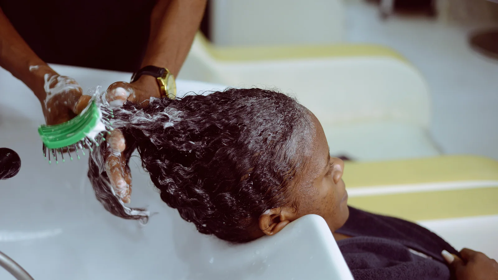 A person washing a woman's hair with foaming shampoo at a salon basin.