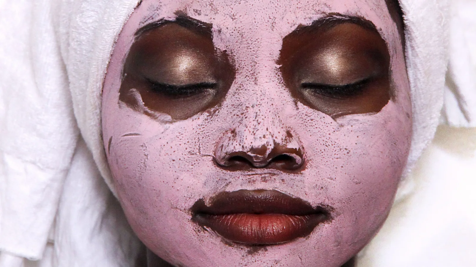 A woman wearing a pale towel and a pink facial mask with her eyes closed.