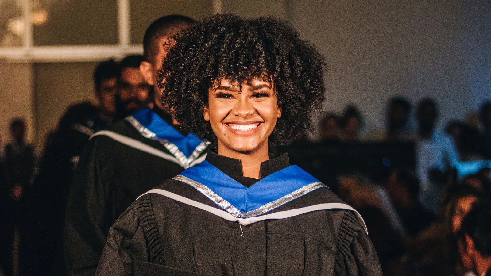 A smiling woman in academic robes standing in front of other graduates.