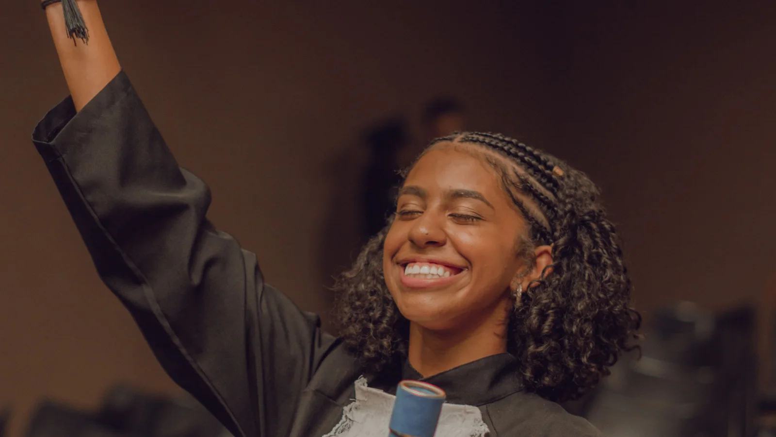 A smiling woman in academic robes lifting a graduation cap.