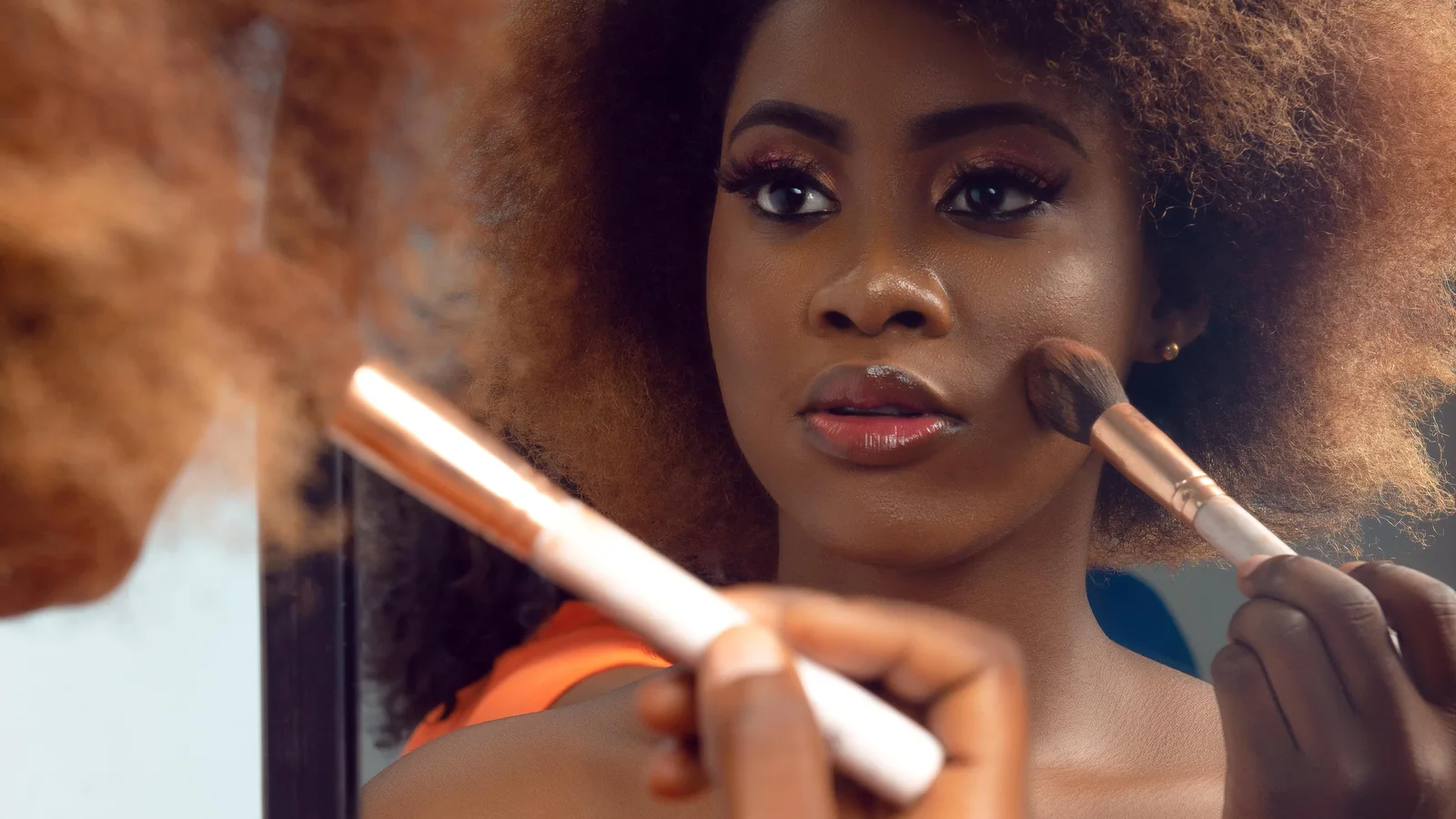 A woman applying make-up with a brush while looking in a wall mirror.