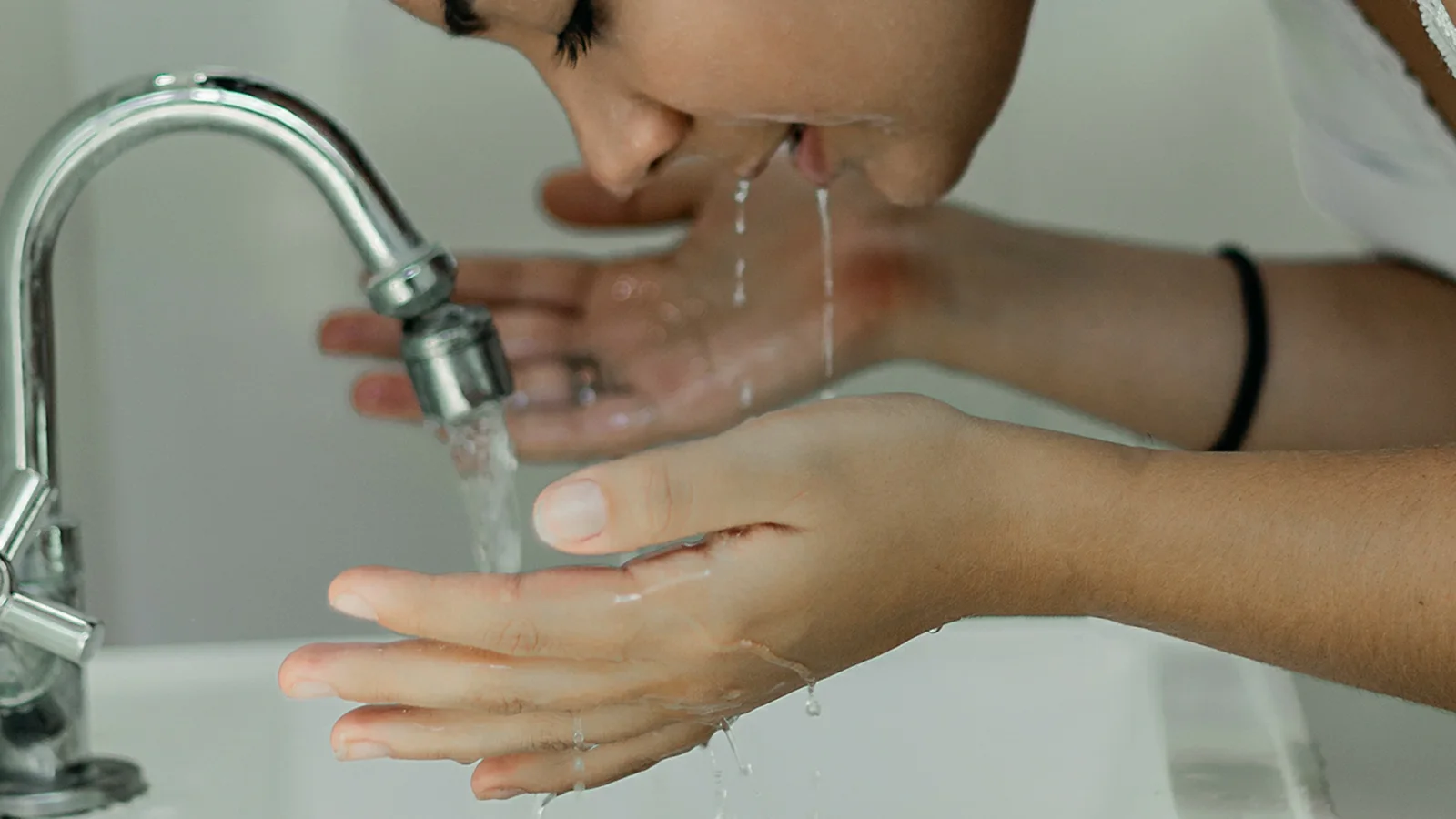 A woman cupping water towards her face at a bathroom sink.