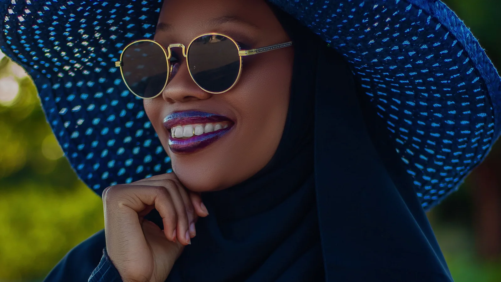 A woman outdoors wearing a wide-brimmed blue hat, sunglasses and a dark head covering.