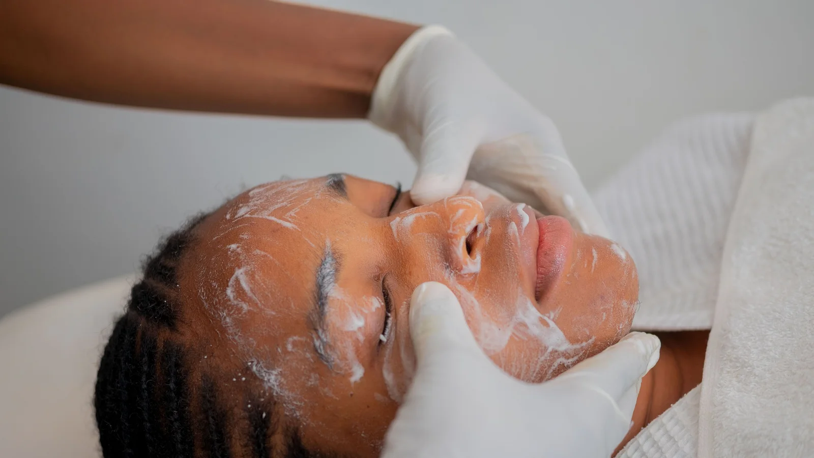 Gloved hands gently supporting a reclining woman's cheeks during a facial.