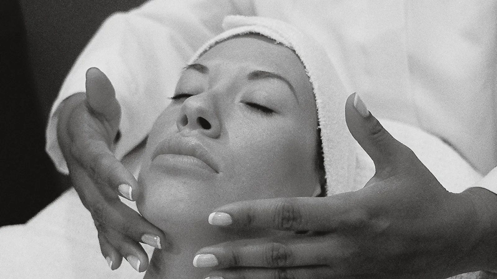 Black-and-white photograph of hands supporting a woman's face during a facial massage.