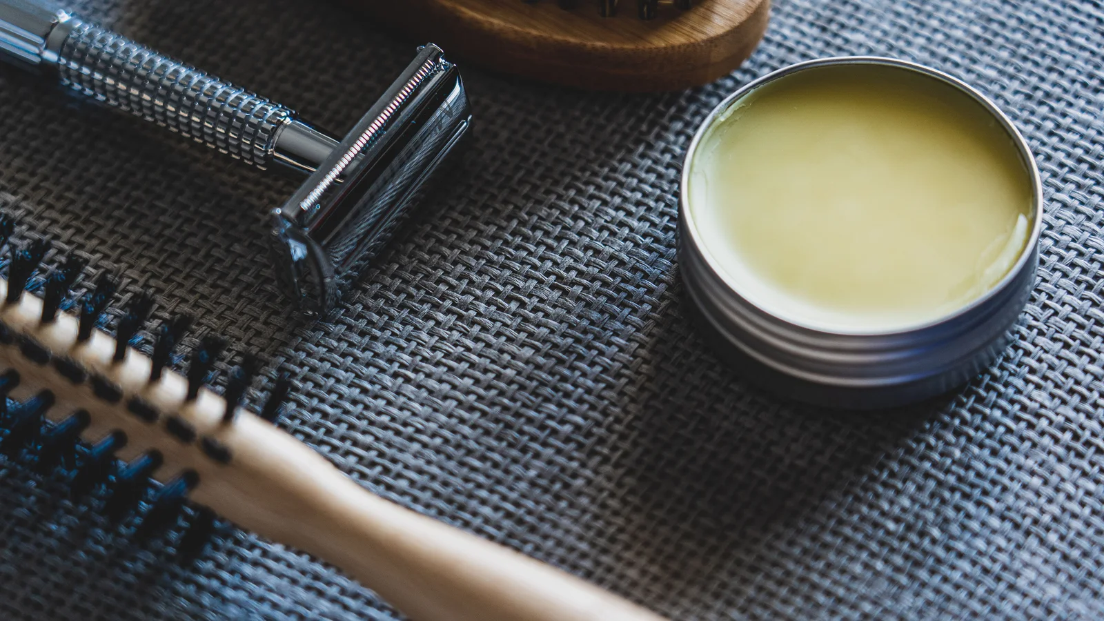 A razor, brush and open grooming-product tin on textured fabric.