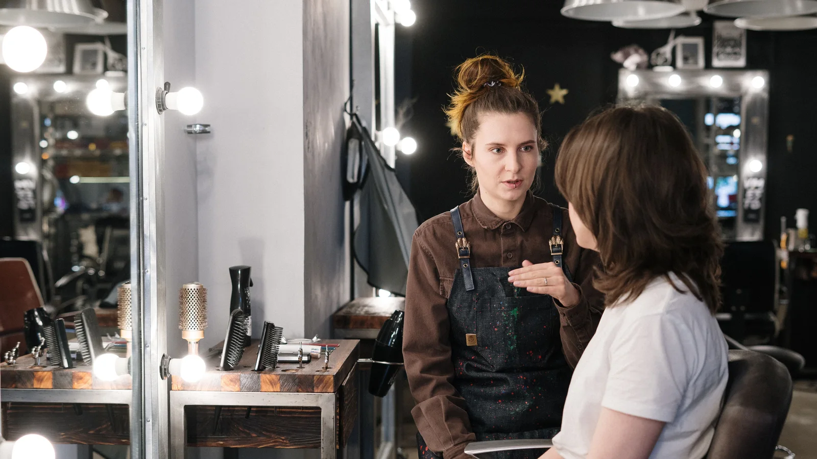 Two women talking while seated beside a salon mirror.