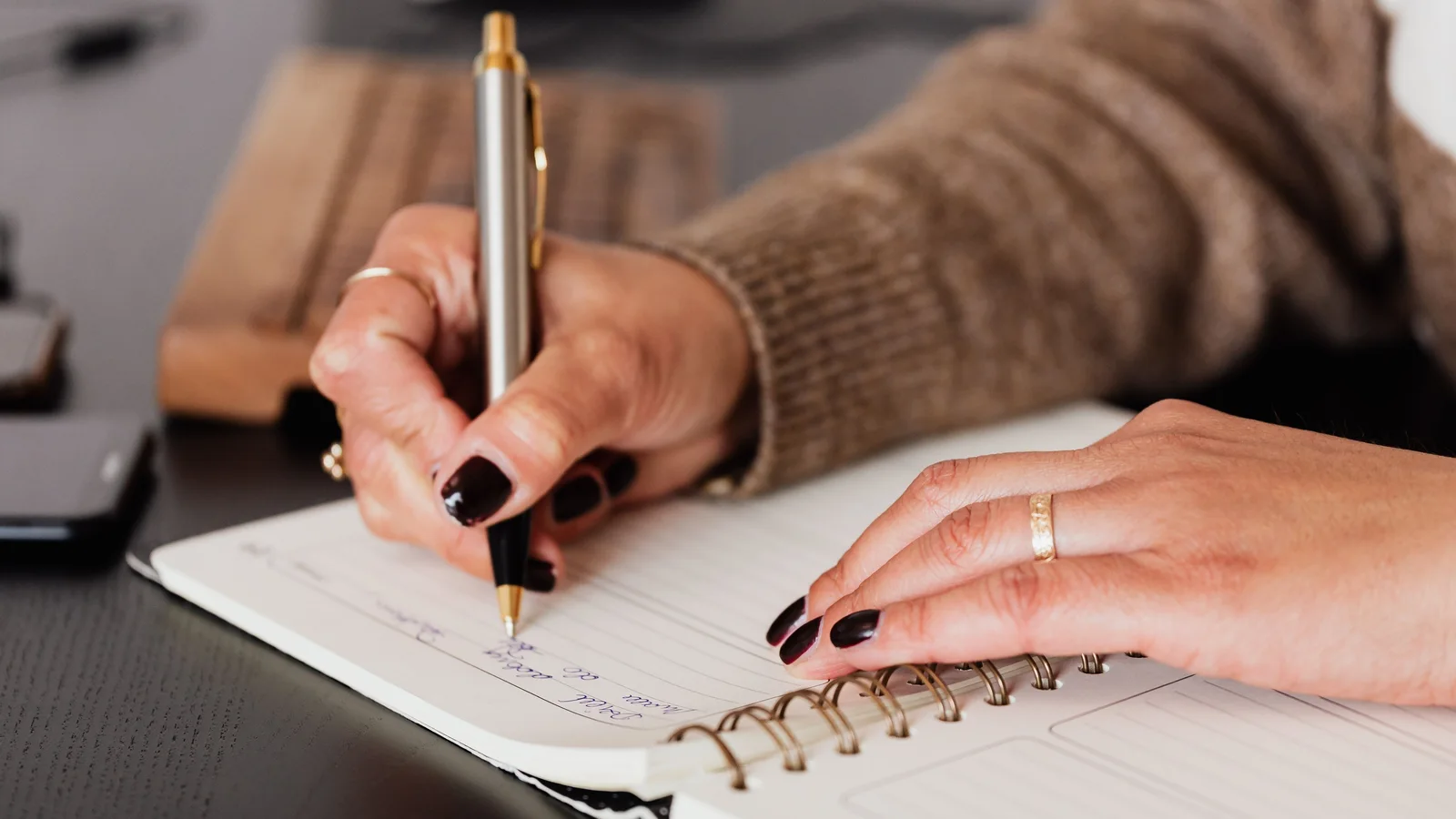 Hands writing in a notebook beside a phone and keyboard.