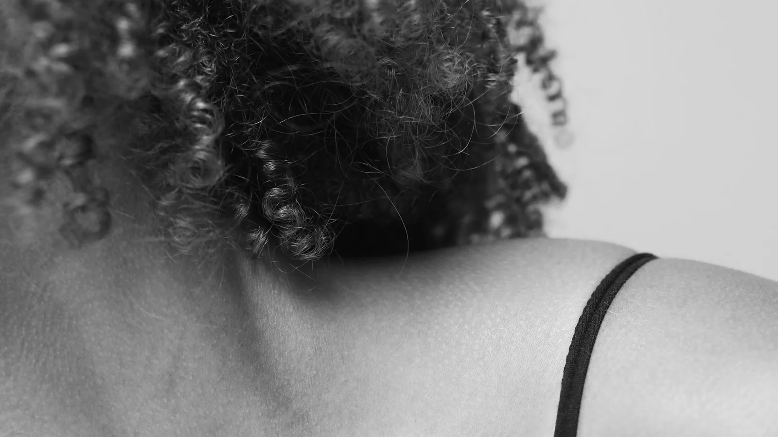 Black-and-white close-up of a shoulder and a narrow clothing strap beside curly hair.