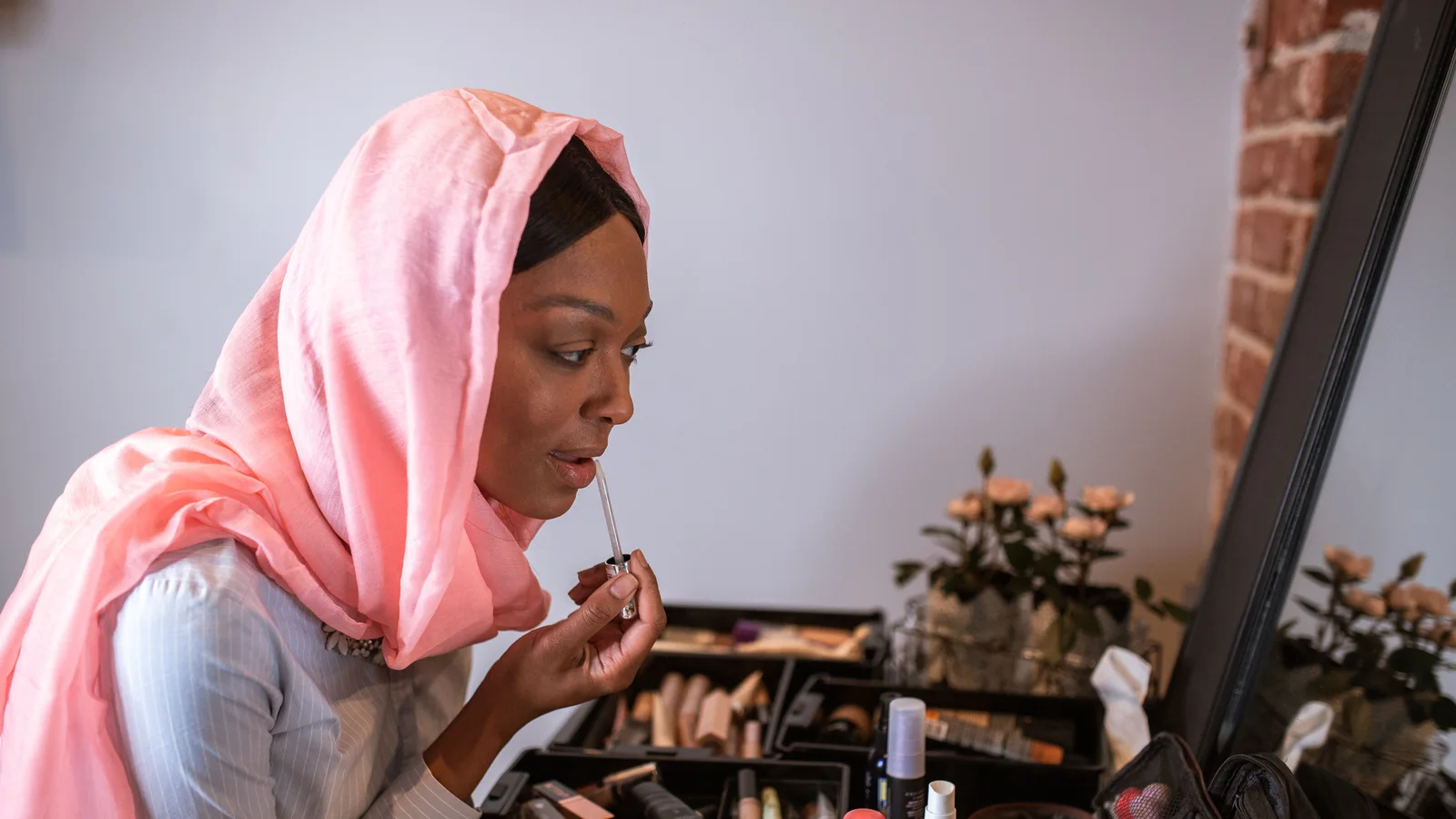 A woman in a pink head covering using a small mirror at a table of make-up.