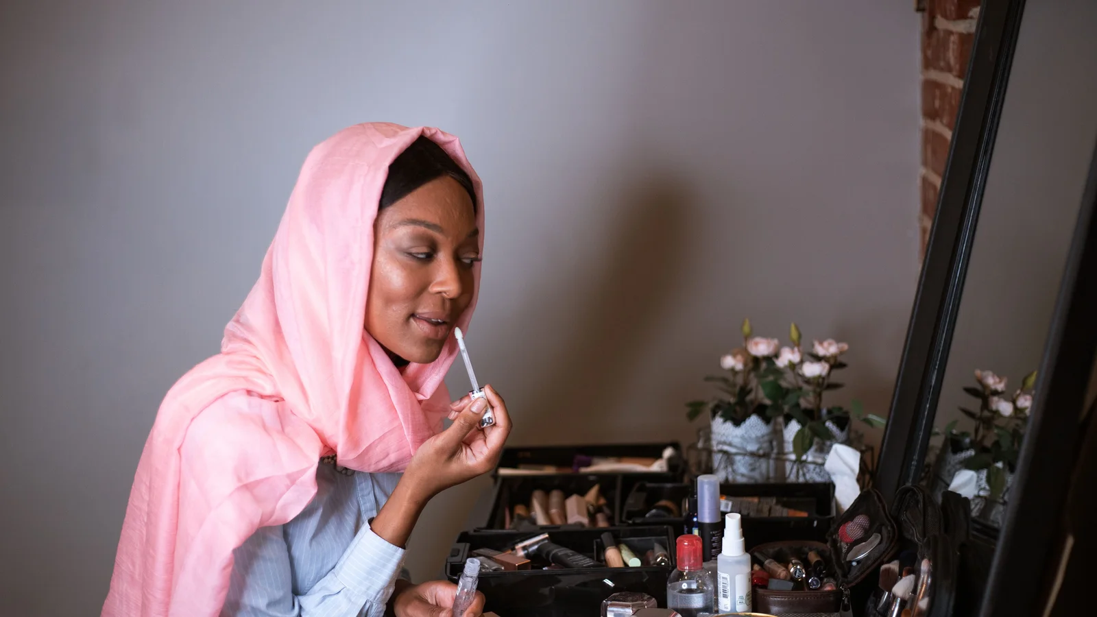 A woman wearing a pink head covering applying make-up at a table of cosmetics.