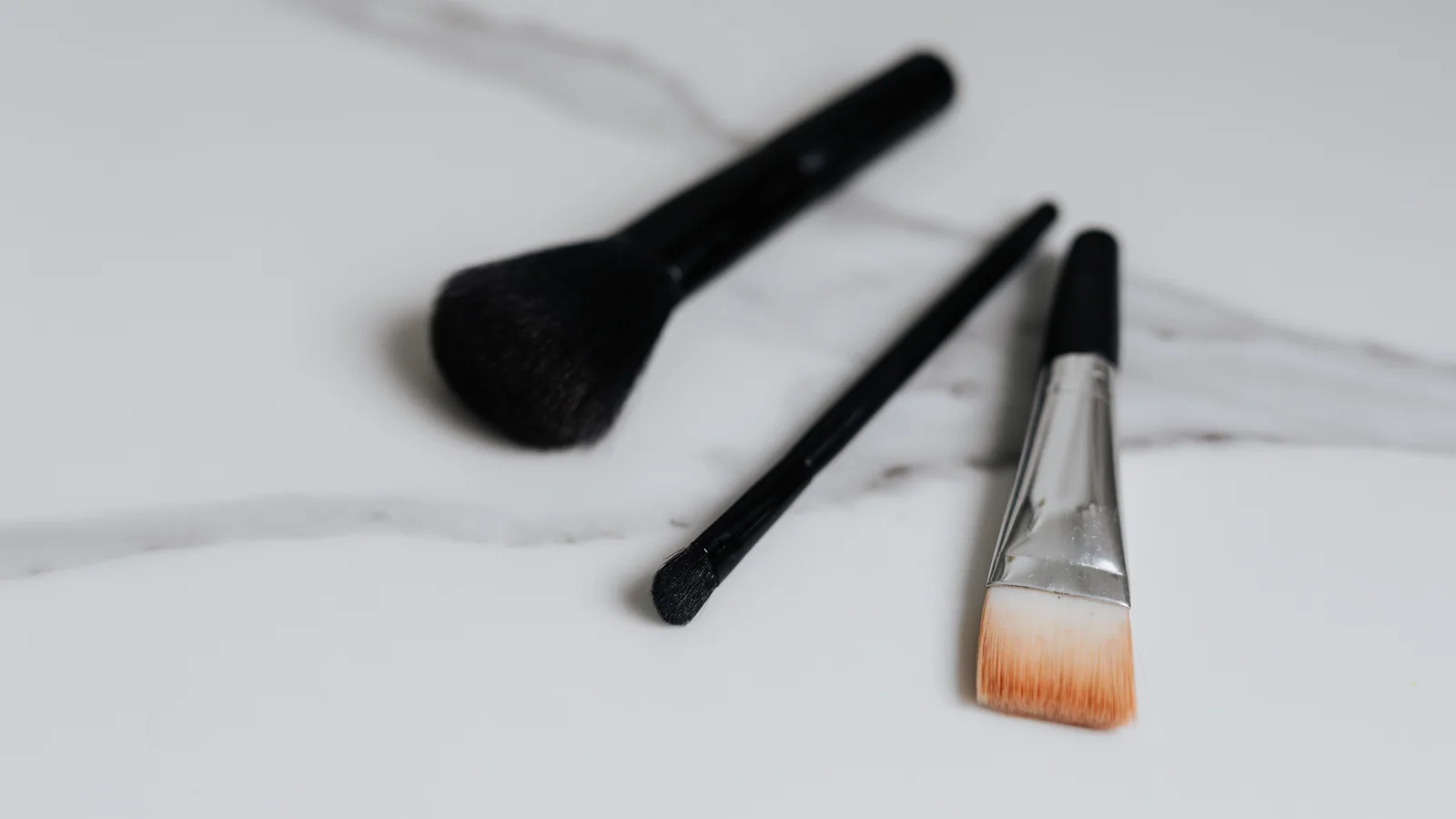 Three make-up brushes lying on a pale marble surface.