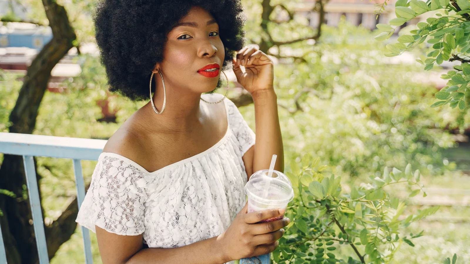 A woman with an afro and red lipstick seated outdoors holding a small glass.