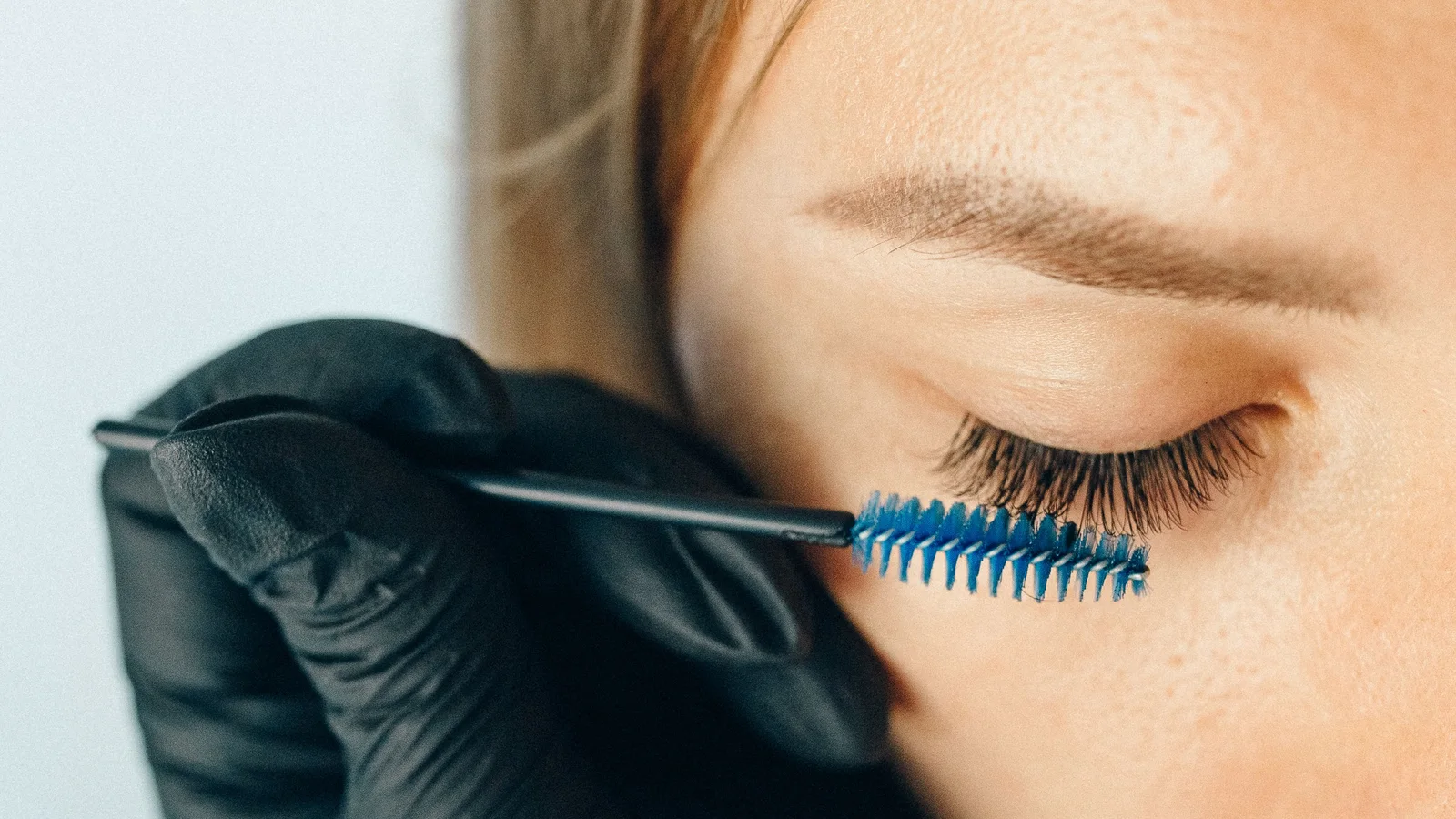 A gloved hand brushing a woman's eyelashes with a blue spoolie.