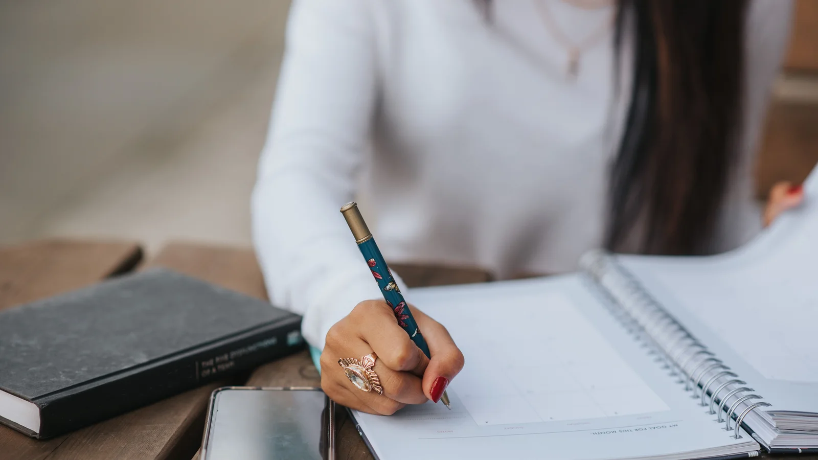 A hand writing in an open notebook beside a phone and a closed diary.