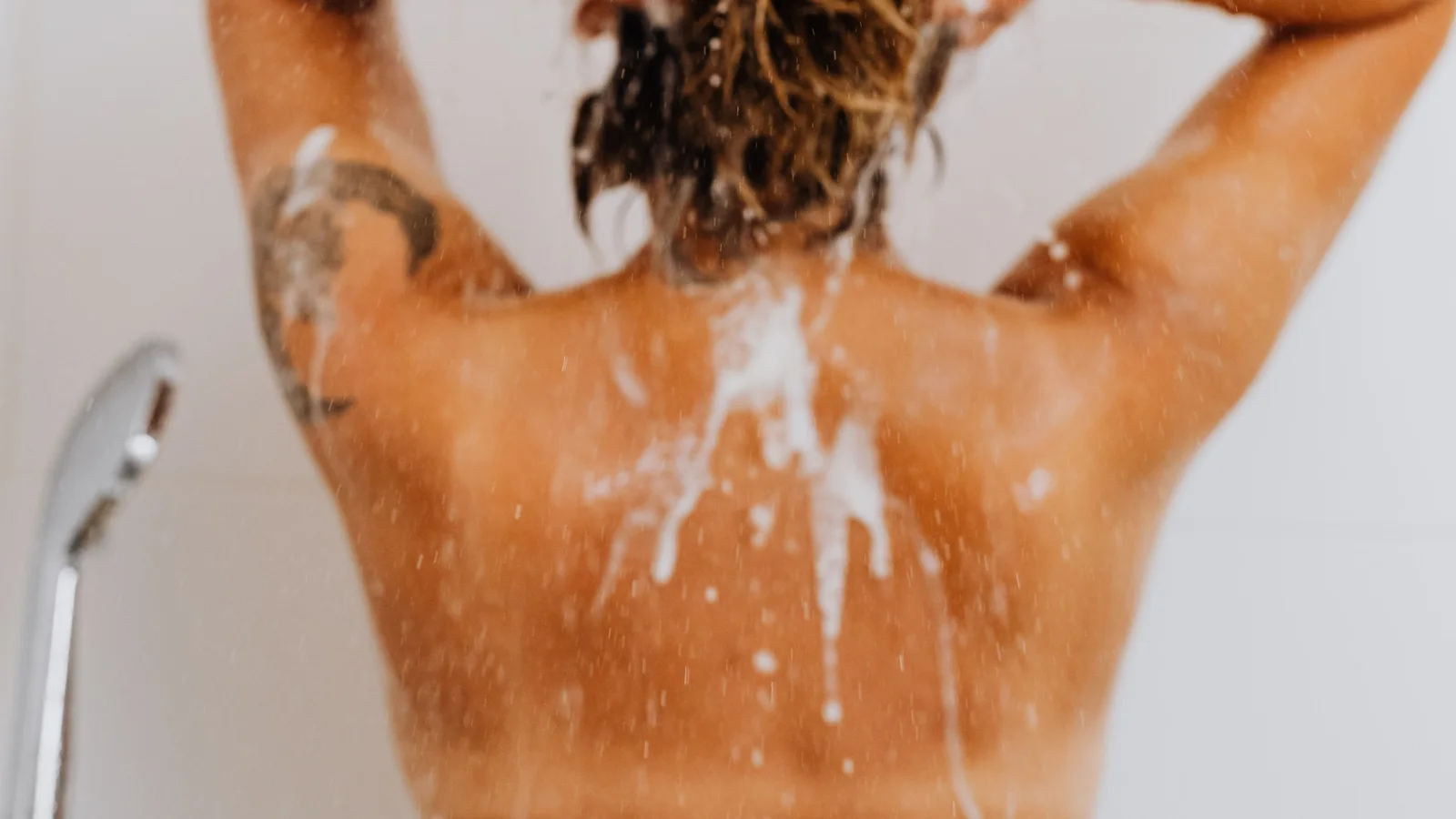 A woman washing her hair under a shower with foam visible on her hands and back.