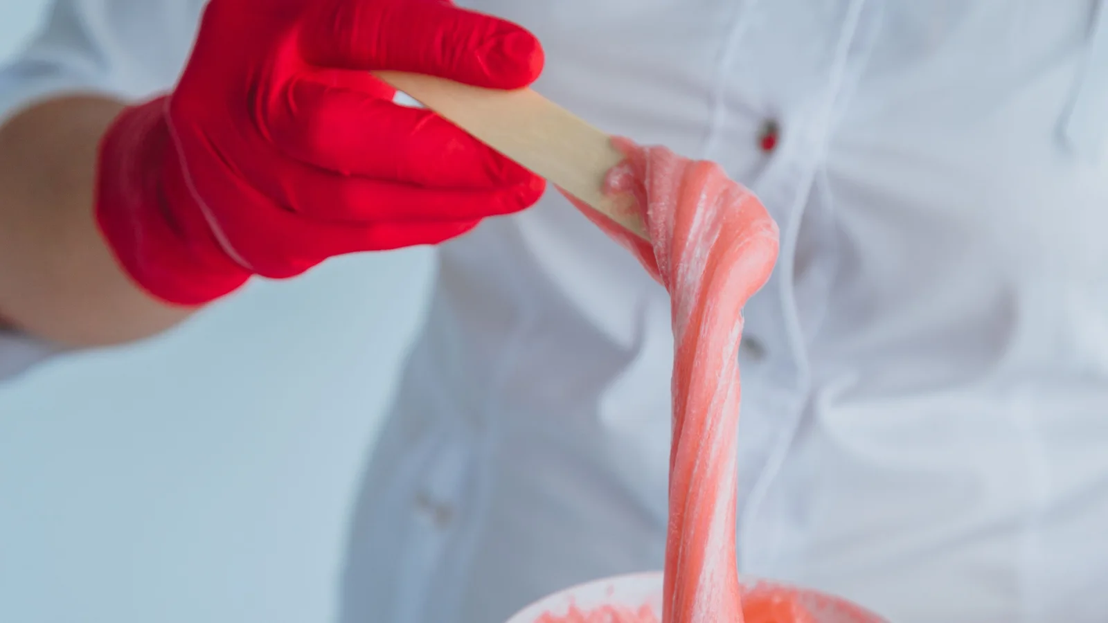 A gloved hand stretching pink sugaring paste from a container with a wooden spatula.