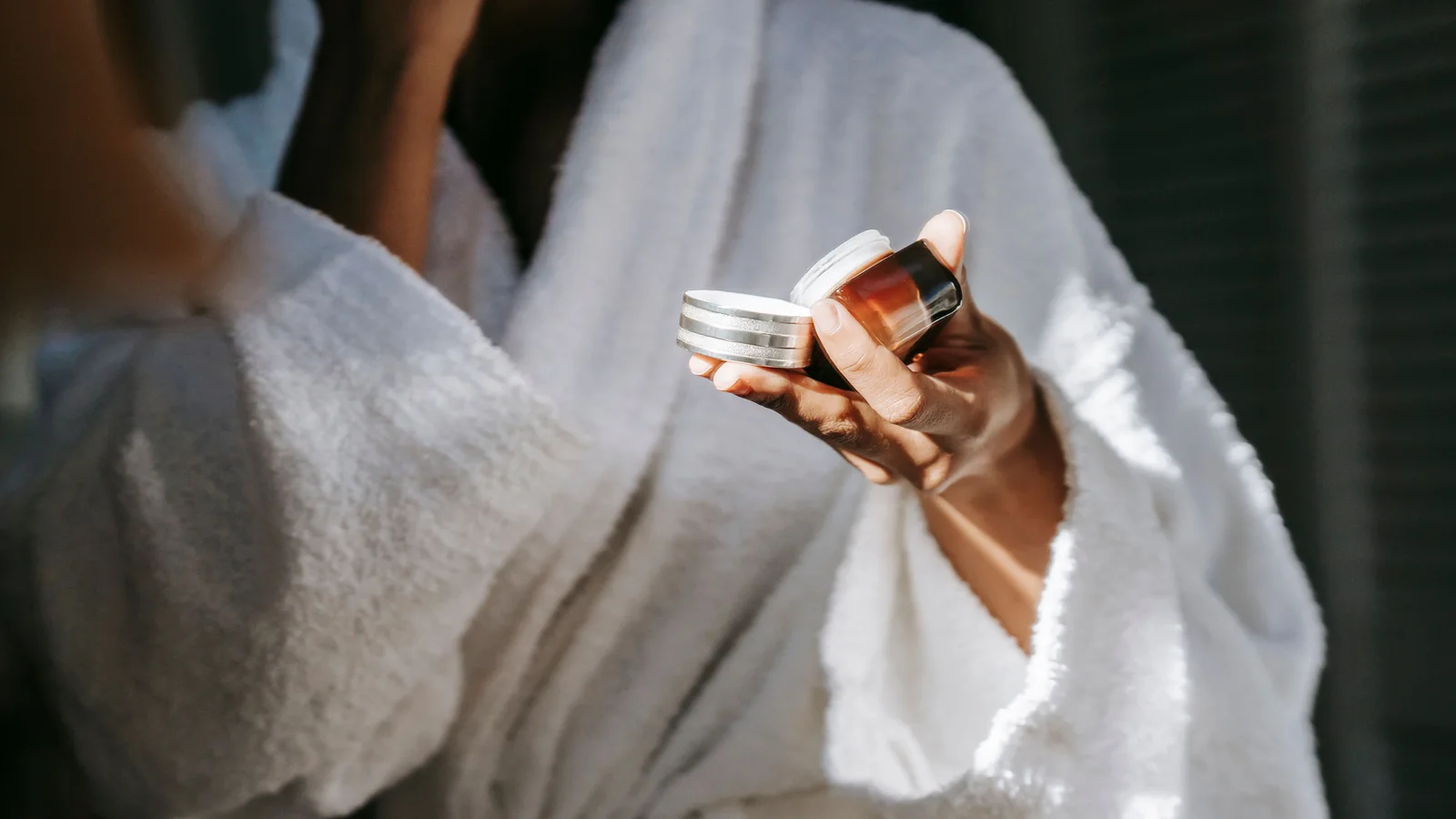 A woman in a white robe holding a cosmetic jar and touching her cheek.