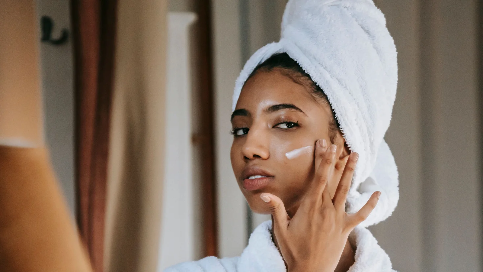 A woman looking into a mirror while touching cream to one cheek.