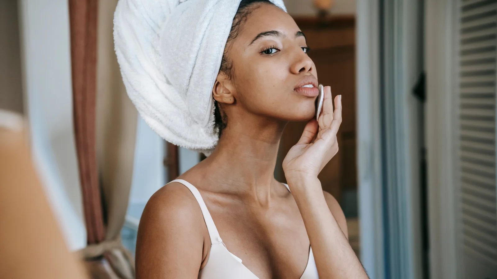 A woman using a cotton pad on her face beside a mirror.