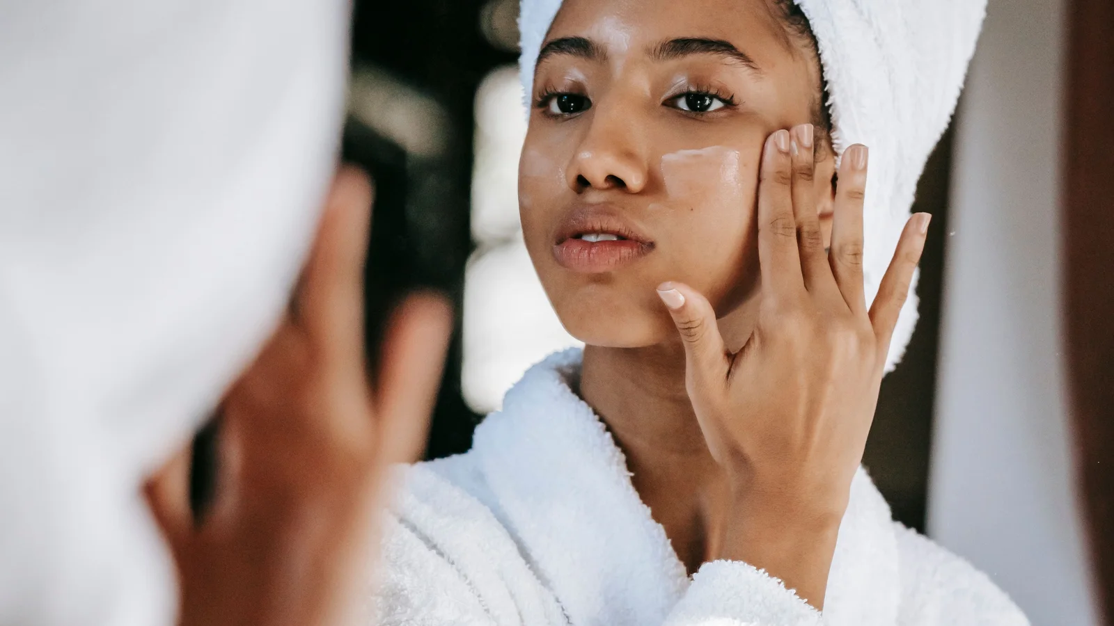 A woman in a white robe applying cream beside a bathroom mirror.