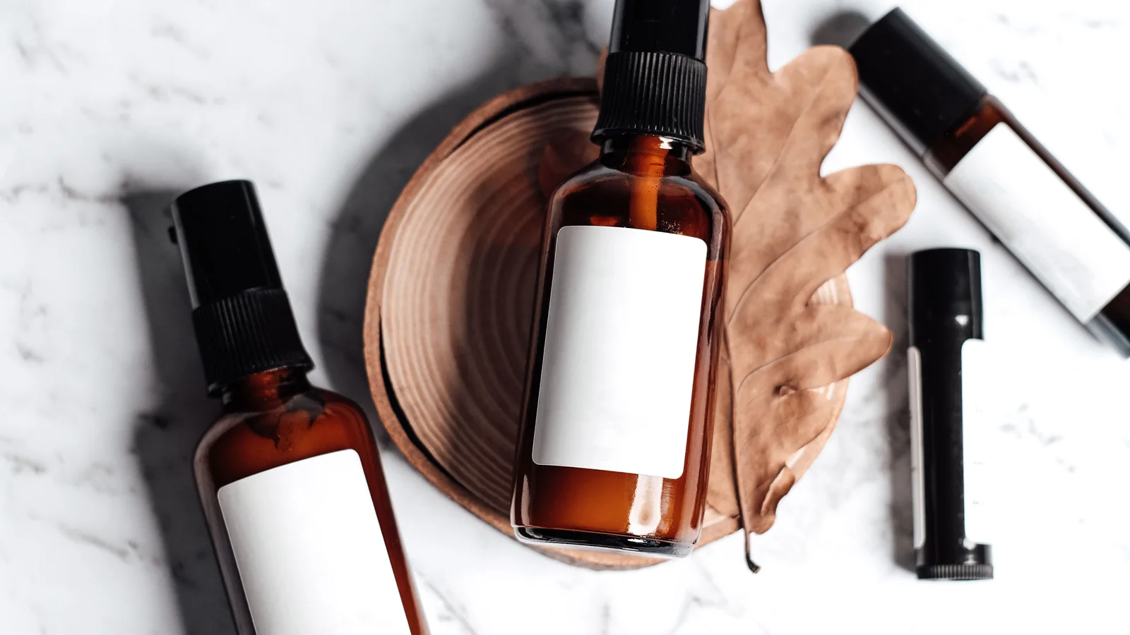 Amber cosmetic bottles with blank labels beside a dry leaf on marble.