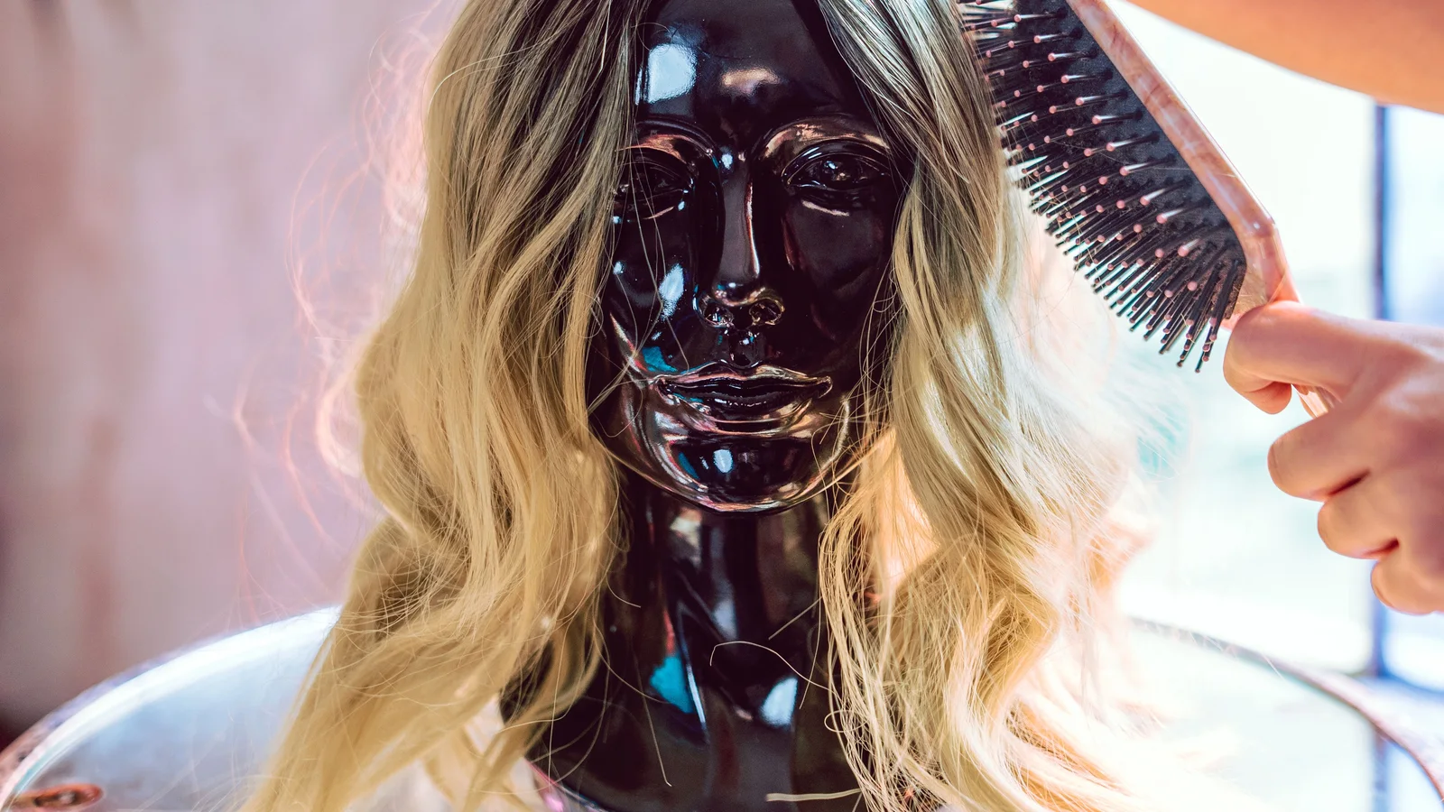 A wig on a mannequin stand being brushed beside a window.